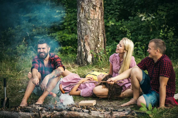 Två par camping i skogen. Killar i röda lumberjack skjortor matlagning korv över eld. Ler vänner avkopplande runt lägerelden på kvällen — Stockfoto