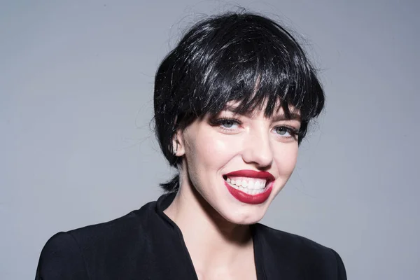 Chica en la cara sonriente feliz lleva chaqueta formal negro. Señora de peluca negra con maquillaje sobre fondo gris. Hermoso concepto de sonrisa. Mujer con atractivos labios rojos mira a la cámara . — Foto de Stock