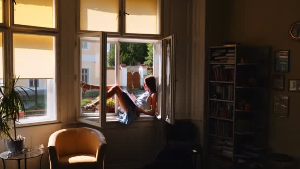 Молодая девушка сидит на окне и мечтает — стоковое видео
