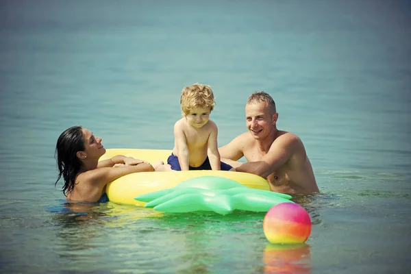 Семья проводит время вместе и веселится. Милый мальчик сидит на надувном матрасе с родителями в форме ананаса в океане, море. Семейный отдых. Отец и мать возле матраса плавают с сыном . — стоковое фото