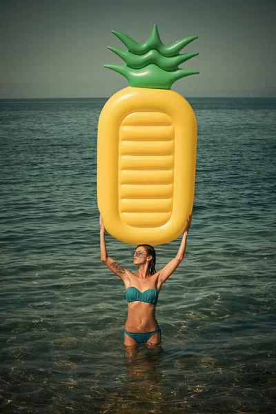 Woman holds above herself air mattress pineapple shaped, sea or ocean on background. Summer vacation concept. Lady with air mattress stand in water, wearing stylish bikini and sunglasses.
