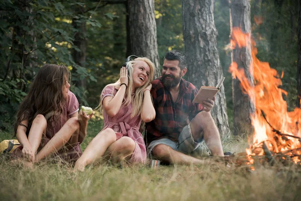 Φίλοι χαλαρώστε στη φωτιά φλόγα με σπινθήρες σε στυλ vintage. Άνθρωποι κάμπινγκ στην πυρκαγιά στο δάσος. Γυναίκες και γενειοφόρος άνδρας με campfire. Κατανάλωση τροφίμων, διαβάζοντας το βιβλίο και την ψυχαγωγία. Καλοκαιρινές διακοπές έννοια — Φωτογραφία Αρχείου