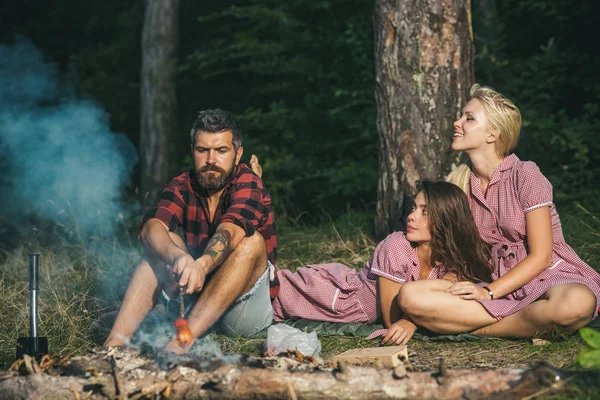 Freunde zelten im Wald. zwei verspielte Mädchen in Vintage-Kleidern, die Holzfäller anschauen. Typ mit ernstem Blick sitzt am Lagerfeuer und fliegt Würstchen — Stockfoto