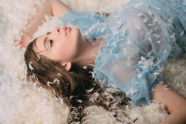 女士在透明的蓝色睡衣躺在床上的羽毛堆 长头发的妇女在柔软的睡衣放松 女孩在神秘的脸上躺在床上布满羽毛和绒毛 香水概念 — 图库照片