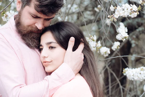 Couple in love spend time in spring garden, branches with flowers on background. Man and woman hugs in blooming garden on spring day. Couple hugs near blooming trees. Spring date concept.