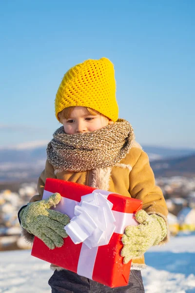 クリスマスプレゼントを持って幸せな子供。クリスマス休暇だギフト付きの赤い箱を持っている編んだ黄色い帽子のかわいい未就学児の男の子。美しい冬の自然。明けましておめでとうございます。. — ストック写真