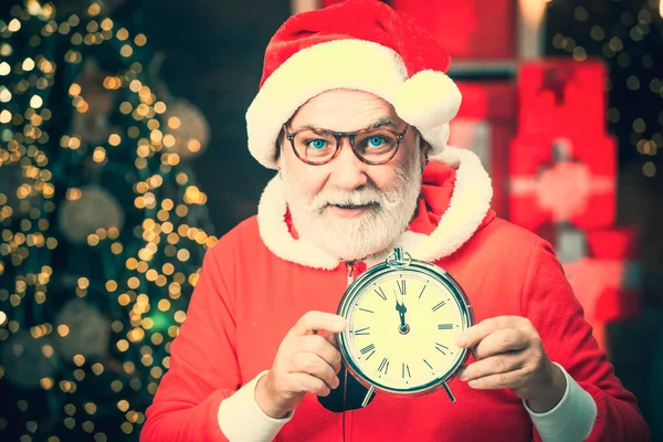 Φωτογραφία από το ρολόι που δείχνει πέντε λεπτά πριν τα μεσάνυχτα. Χαρούμενος Άγιος Βασίλης κρατώντας ξυπνητήρι στο τζάκι και φόντο χριστουγεννιάτικο δέντρο. Ευχετήρια χριστουγεννιάτικη κάρτα. — Φωτογραφία Αρχείου
