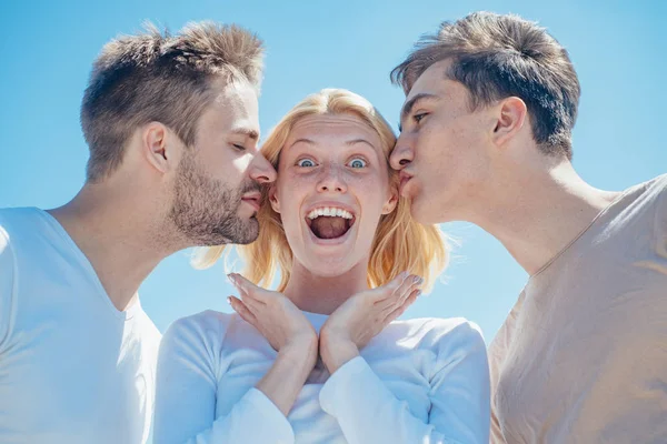 Ξανθιά, χαρούμενη κοπέλα, να δέχεται φιλιά από δύο όμορφα αγόρια. Γοητευτική γυναίκα στέκεται ανάμεσα σε δύο φίλους. Η ιδέα των καλύτερων φίλων. Ομάδα φίλων στο καθαρό φόντο του ουρανού. — Φωτογραφία Αρχείου