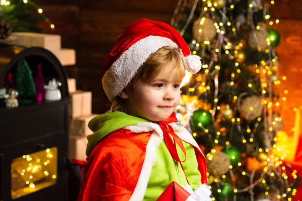 Criança feliz em chapéu de Pai Natal no fundo da árvore de Natal. Tempo de Natal. Menino bonito criança alegre humor jogar perto da árvore de Natal. Presentes para férias de inverno perto do lugar de fogo . — Fotografia de Stock