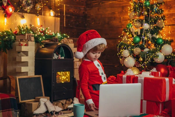 İnternette sörf yapan akıllı çocuk. Noel Baba küçük yardımcısı. Küçük çocuk Noel Baba şapkası ve kostümü. Noel ağacının yanında dizüstü bilgisayarı olan bir çocuk. İnternetten noel hediyesi al. Noel alışverişi konsepti. Hediye servisi — Stok fotoğraf