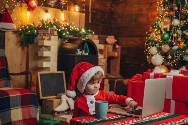 サンタはノートパソコンの赤ちゃんを助ける。新年の明るいインテリア。クリスマスツリー屋内だクリスマスツリーの子供とクリスマスの前夜の暖炉。クリスマスだクリスマスの属性. — ストック写真