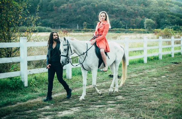 Couple in love walking with horse on countryside. Romantic love story of sensual couple. Woman riding grey arabian horse in pink dress. Handsome bearded man driving horse for a bridle and looking at — Stock Photo, Image