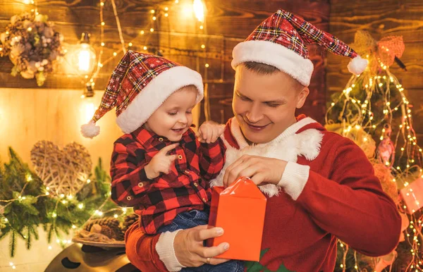 サンタの帽子の笑顔の父と彼の息子はクリスマスプレゼントボックスを開きます。新年の木と火の場所を背景に。クリスマスだ子供の頃の記憶クリスマスの子供の夢. — ストック写真