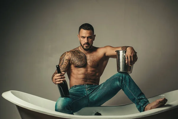 Knappe baardloze man in jeans met sexy lichaam in de badkamer. Seksuele macho man in bad. Sterke gespierde tattoed man met champagne fles en poseren in de badkamer. — Stockfoto