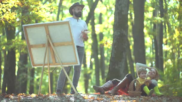 Муж художник рисует картину на осенней природе. Улыбающаяся пара с ребёнком, гуляющая по осеннему естественному фону. Веселая семья проводит пикник, расслабляясь вместе на желтой осенней природе в парке . — стоковое видео