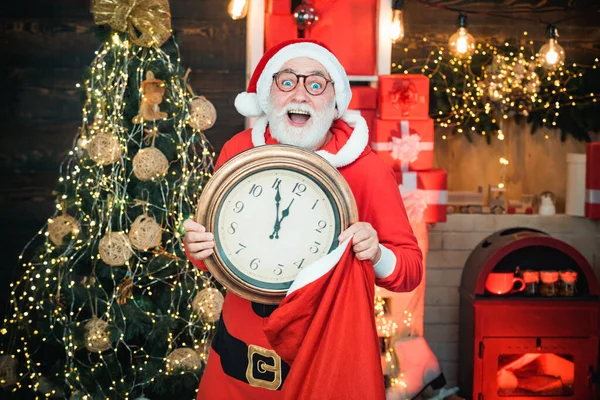 Weihnachtsmann hält Uhr mit Countdown zu Weihnachten oder Neujahr Weihnachtsmann in Holzhaus Interieur zeigt die Zeit auf einer Uhr. — Stockfoto