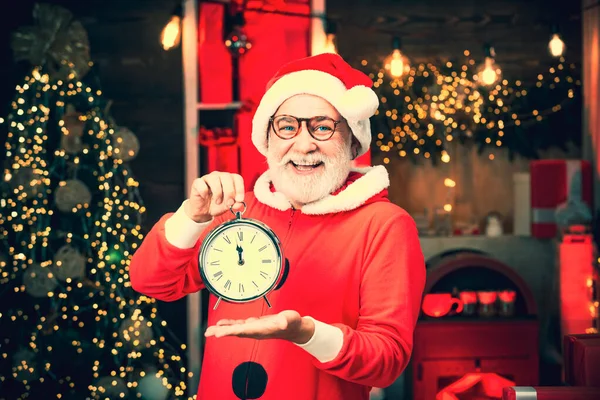 Der Weihnachtsmann macht ein lustiges Gesicht und hält eine Uhr, die fünf Minuten vor Mitternacht zeigt. Der Weihnachtsmann hält einen Wecker vor dem Hintergrund des Weihnachtsbaums. Der Weihnachtsmann zeigt auf der Uhr. — Stockfoto