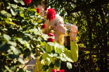 Bir bardak su içen, gözlerini kapayan, sağlıklı bir yaşam tarzını gösteren genç ve çekici bir kadının portresi. Güller arasında kızların yüzünü kapatın. Nem, dengeli
