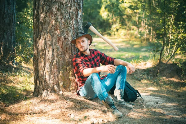 O lenhador a trabalhar numa floresta. Bonito Woodworkers lenhador xadrez camisa segurando o machado em fundo natureza verde. Um jovem bonito com barba carrega uma árvore . — Fotografia de Stock