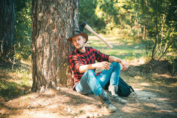 The Lumberjack working in a forest. Handsome Woodworkers lumberjack plaid shirt holding the axe on green nature background. A handsome young man with a beard carries a tree.
