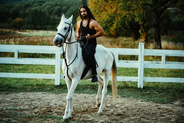 Homem atraente sentado em cavalo branco no rancho no outono. Um jovem bonito sentado no garanhão do lado do campo. Homem equestre em seu cavalo montando na natureza. — Fotografia de Stock