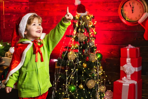Lindo niño de Santa muestra el pulgar hacia arriba en decorado fondo del árbol de Año Nuevo. Montaña de regalo y cajas de regalo. Ayudante de Santa con regalos mágicos de Navidad . — Foto de Stock