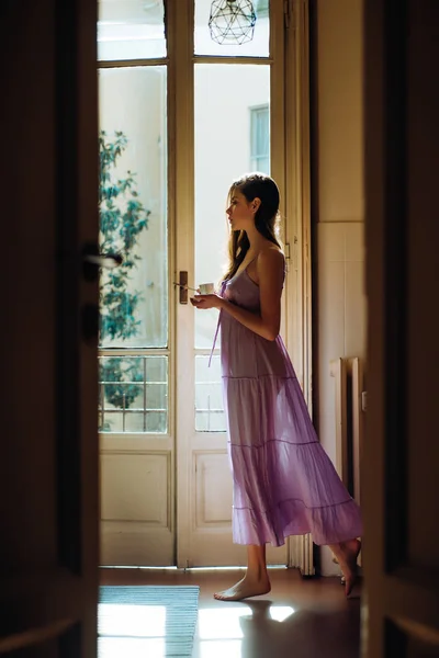 Trzyma kubek, który tańczy. Dziewczyna jest ubrana w koszulę nocną. Balkon z widokiem na dom. Portret pięknej dziewczyny z długimi włosami. Wschód słońca o świcie. — Zdjęcie stockowe