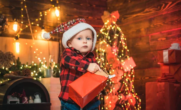 Frohe Weihnachten und frohe Feiertage. Lustiges Baby öffnet ein Weihnachtsgeschenk in seinen Händen auf dem weihnachtlichen Hintergrund. Weihnachtsbaum. Glückliches Kind. — Stockfoto