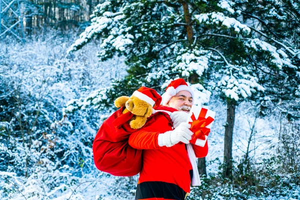 冬の森を歩く赤い衣装を着たサンタクロース。クリスマスプレゼントをサンタクロースは雪に覆われたフィールドで冬に贈り物の袋を持って行く. — ストック写真