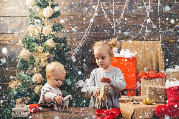 Niedliches kleines Mädchen schmückt den Weihnachtsbaum drinnen. Frohe Weihnachten. Porträt Kind mit Geschenk auf Holzgrund. Lächelndes Kind lugt hinter Weihnachtsbaum im Wohnzimmer hervor. — Stockfoto