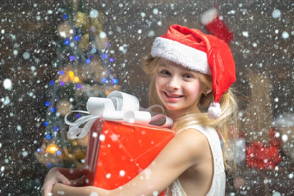 Roztomilá holčička je zdobení vánočního stromku uvnitř. Šťastné děti. Šťastné malé teenager v Santa čepice s současnosti mají Vánoce. Portrét kid s darem na dřevěné pozadí. — Stock fotografie