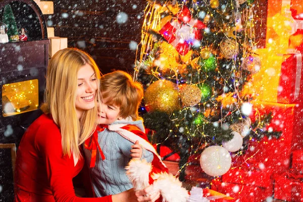 Alegre sorridente mãe abraços com seu filho querido na árvore de Natal e apresenta fundo. Criança pequena feliz brincar com decorações de Natal. Atributos de Natal . — Fotografia de Stock