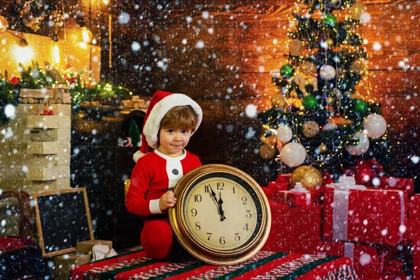 Navidad o Año Nuevo. Lindo niño en sombrero de Santa Claus sosteniendo despertador rojo con el pulgar hacia arriba. Doce Oclock medianoche en estilo retro. Decoración de Navidad. muestra del reloj justo antes de Año Nuevo . — Foto de Stock