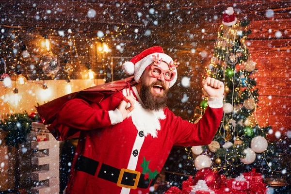 Crazy, funny Hipster Santa. Bearded man having fun near Christmas tree indoors. New year Christmas concept. Handsome man wearing in Christmas dress. — Stock Photo, Image