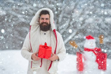 Noel hazırlığı. Kırmızı hediye kutusu olan komik sakallı adam kardan adam yapıyor. Kış tatili ve insanların konsepti. Noel ve Noel 'i kutluyoruz.