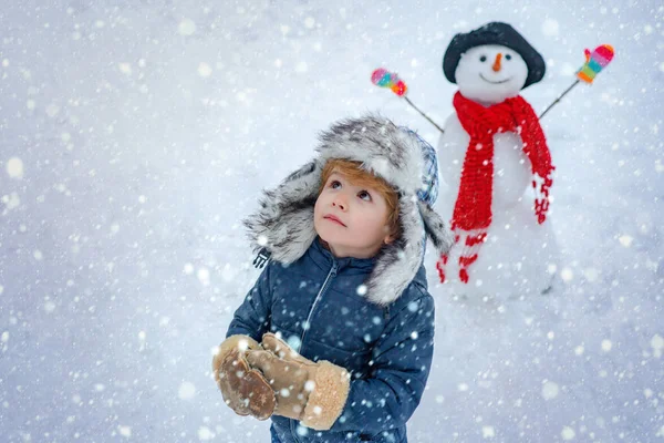 Зимнее утро. Милый маленький мальчик на снежном поле под открытым небом. Веселый ребенок веселится со снеговиком в Зимнем парке. — стоковое фото