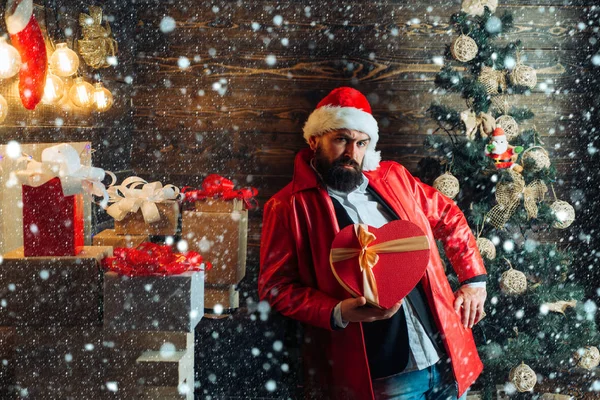 Christmas man in snow. Bearded modern santa wishes Merry christmas and Happy new year. Christmas Celebration, holiday. Christmas tree background. Santa claus with red Christmas gift box. — Stockfoto