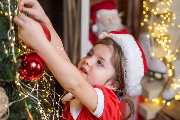Neujahrs- und Weihnachtskindkonzept. Weihnachtskind schmückt Weihnachtsbaum mit Christbaumkugel. Kleines Mädchen mit Weihnachtsmütze schmückt Weihnachtsbaum mit Christbaumkugeln. — Stockfoto