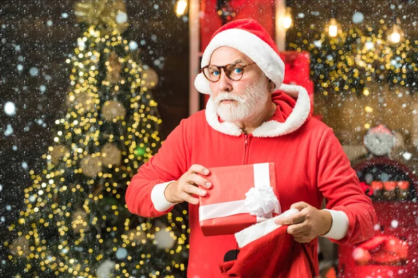 Uomo di Natale sulla neve. Babbo Natale divertente tenere regalo di Natale. Babbo Natale augura buon Natale. Santa nonno con una barba bianca in posa sullo sfondo di legno di Natale . — Foto Stock