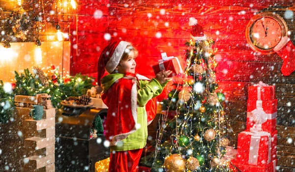 雪の中のクリスマスの子供たち。クリスマスツリーによって幸せな小さな男の子誰かにクリスマスの贈り物を与える。小さな子供はサンタの服を着ている. — ストック写真