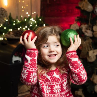 Kar taneleri ve geyikleriyle kırmızı ceketli küçük kız. Tatil konsepti. Kış kazaklı mutlu küçük kız anaokulu öğrencisi elinde kırmızı ve yeşil Noel ağacı topları tutuyor. Noel zamanı.