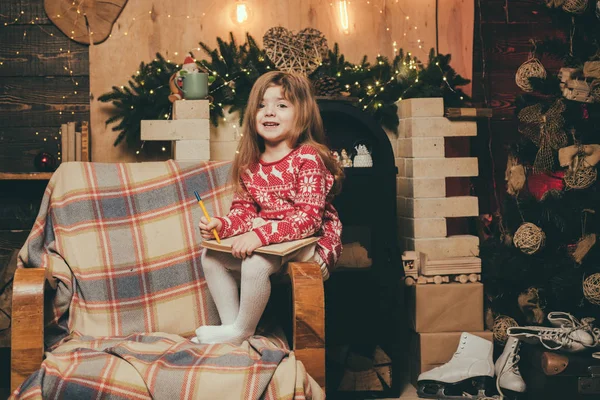 クリスマスの女の子はクリスマスツリーに座って彼女のノートに何かを書いています。屋内でクリスマスツリーの近くで楽しんでいる子供. — ストック写真
