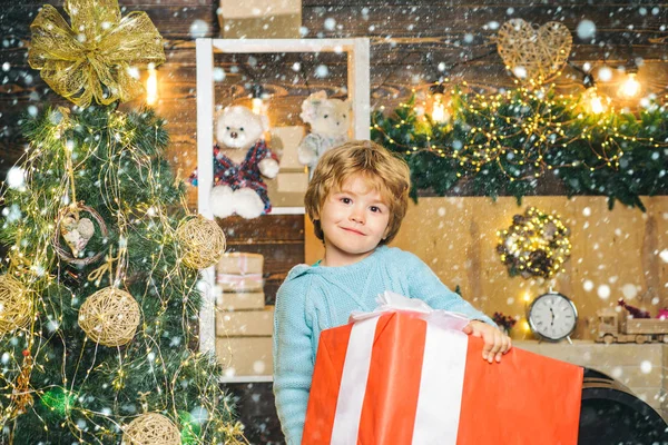 Weihnachtskinder im Schnee. glückliches Kind, das mit beiden Händen eine riesige rote Geschenkschachtel hält. Weihnachtsfeier. fröhliches kleines Kind in Winterkleidung denkt an Weihnachtsmann in der Nähe des Weihnachtsbaums. — Stockfoto