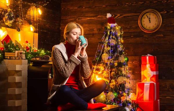 A senhora está bebendo bebida quente e sentada na árvore de Natal. Uma jovem feliz junto à lareira. Inverno, tempo de Natal . — Fotografia de Stock