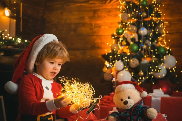 A criança está brincando com a luz de Natal no fundo da árvore de Natal. O miúdo está a mostrar luz Chritmas ao seu brinquedo. Fecha. Boas festas, família . — Fotografia de Stock