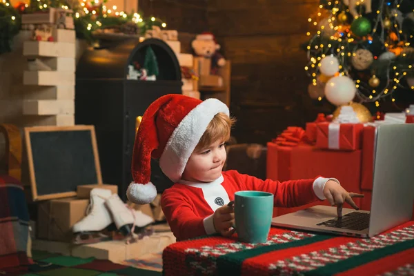 Compras de Navidad en línea para niños. El niño está usando ropa de Santa sentado junto a su portátil. Ayudante de Santa usando portátil. —  Fotos de Stock