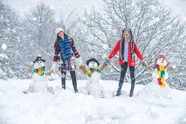 Amigos de Navidad en la nieve. Chicas de invierno con muñeco de nieve. Fiesta de estudiantes y celebración de Navidad. Dos alegres mujeres jóvenes de belleza divirtiéndose con muñeco de nieve en Winter Park. Feliz Navidad y Feliz Año Nuevo . — Foto de Stock