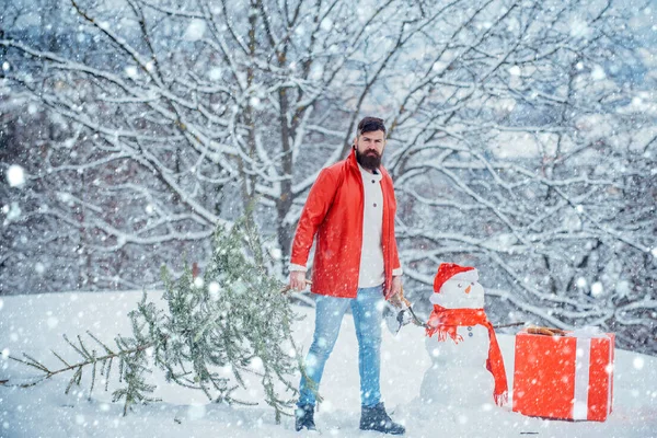 Homme de Noël dans la neige. Drôle Santa homme posant avec hache et arbre de Noël. Un homme barbu porte chez lui un sapin de Noël. Homme bûcheron avec arbre de Noël dans le parc d'hiver . — Photo