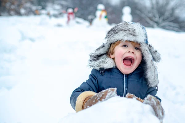白い雪の背景に公園で雪と遊ぶ興奮した子供。寒い冬の公園の冬の子供たち。かわいい子供-冬の肖像画。雪の中で雪だるまと面白い男の子の子供. — ストック写真