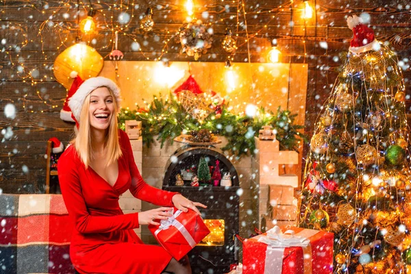 Mulher na neve. Menina engraçada senta-se perto da árvore de Natal, presentes e lareira em roupas de casa. Jovem mulher em vestido elegante sobre fundo interior de Natal . — Fotografia de Stock